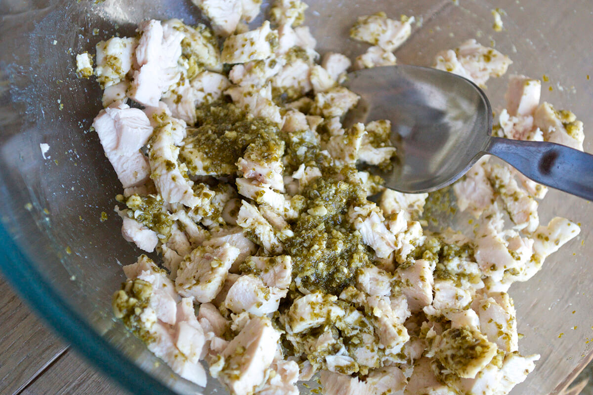 A glass bowl with cooked chicken and pesto being mixed together