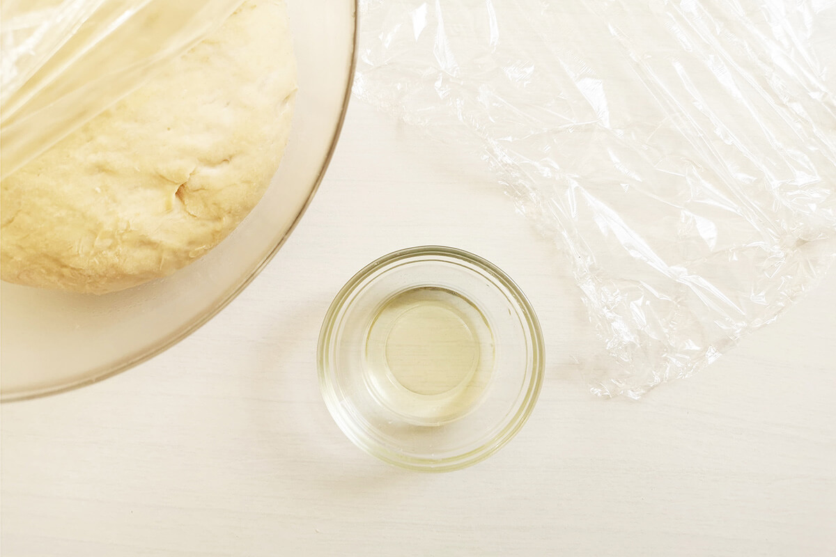 Ball of dough in a glass bowl next to some cling film and a small container of oil