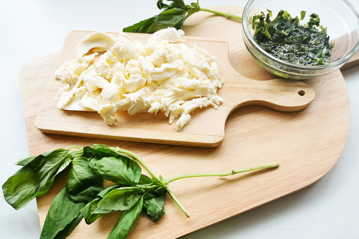 A wooden chopping board with mozzarella, fresh basil leaves and a small container of chopped basil mixed with olive oil