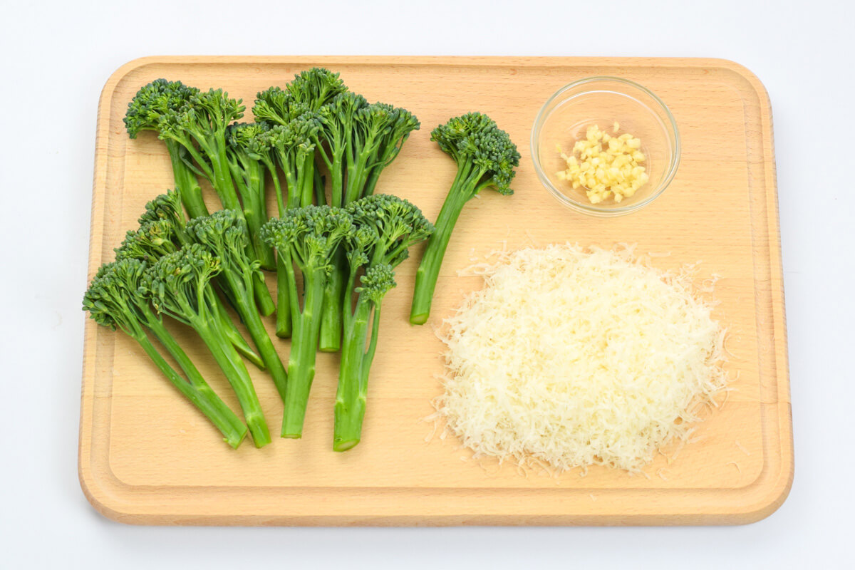 A wooden chopping board with tenderstem broccoli florets, grated parmesan and a small glass bowl of crushed garlic