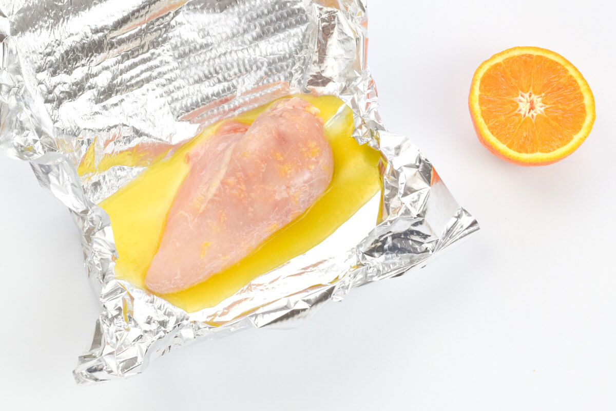 A chicken fillet in foil, with a squeeze of orange juice on it, next to a halved orange