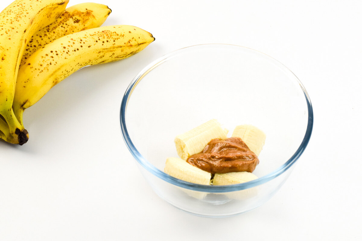A glass bowl with banana and peanut butter, next to 2 ripe bananas