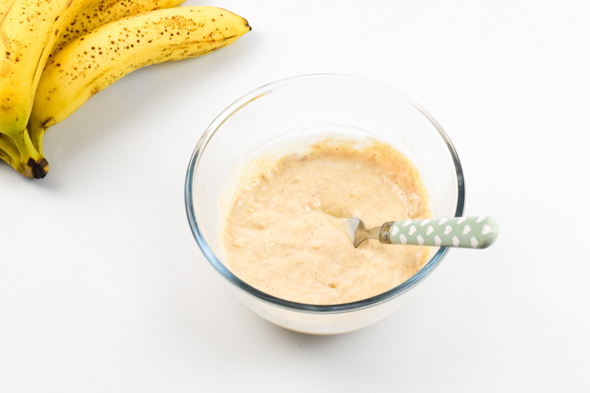 A glass bowl with banana and peanut butter mashed with yoghurt, next to 2 ripe bananas