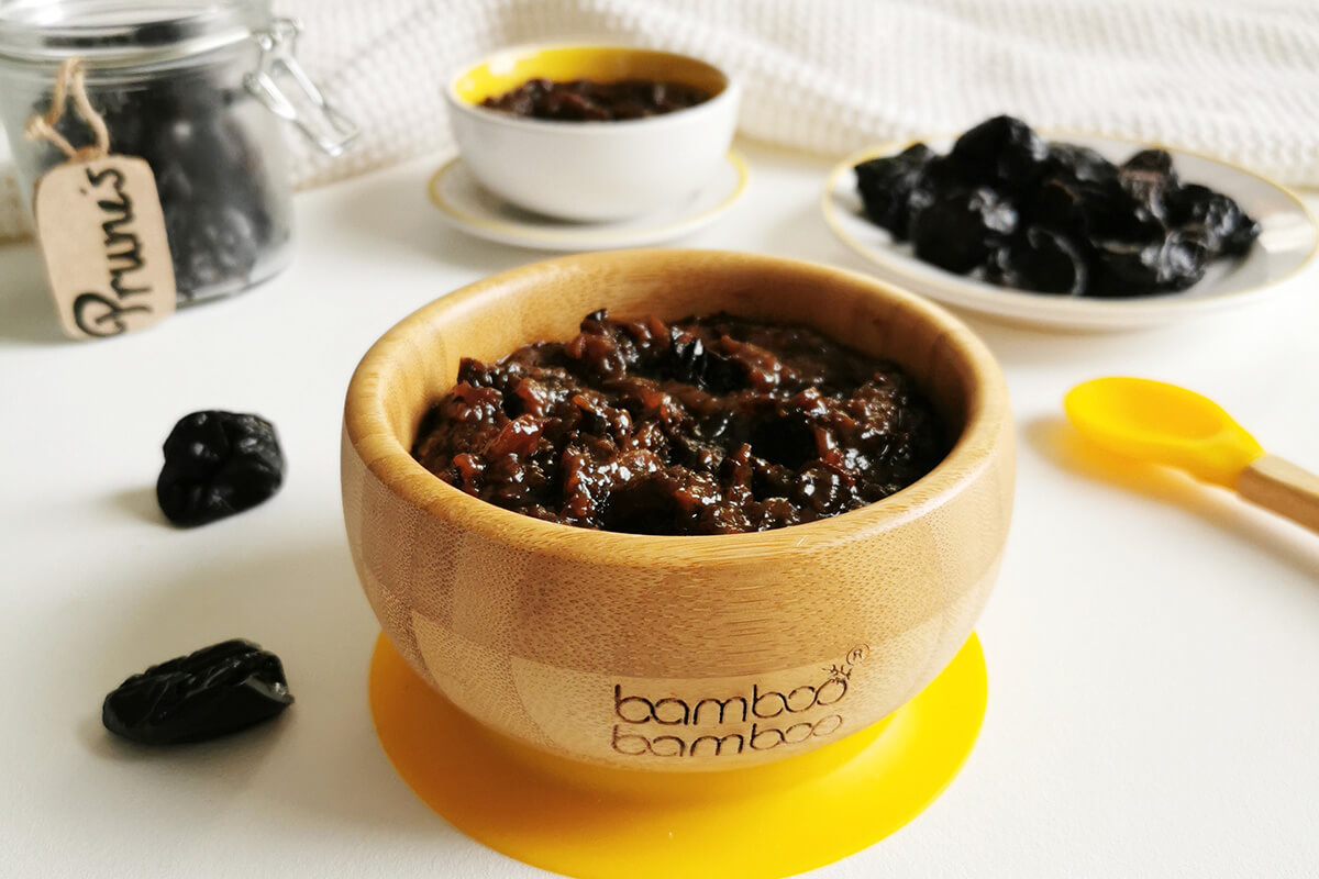 A small bowl of prune puree with some prunes around it and next to a jar of prunes, a small plate of prunes