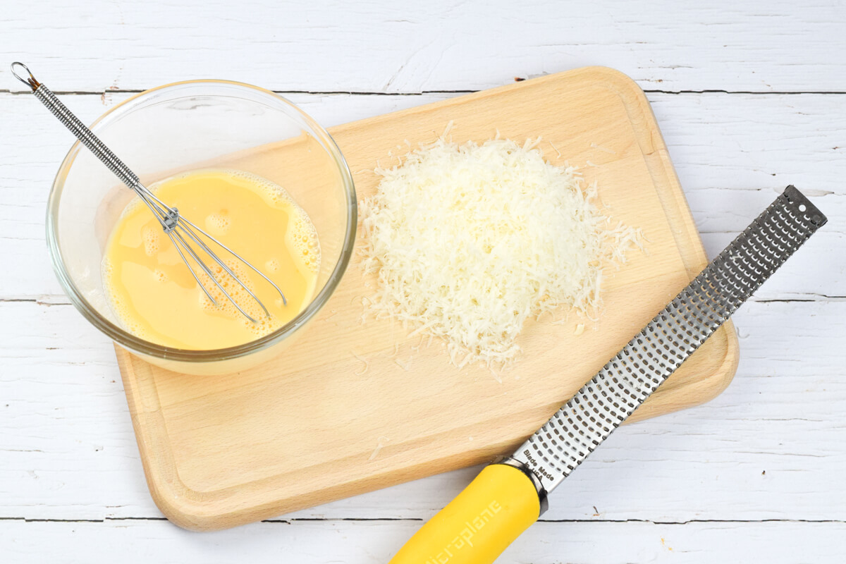 A wooden chopping board with grated parmesan next to a glass bowl of whisked eggs