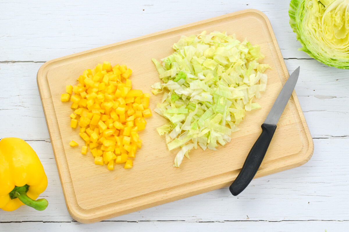 A wooden chopping board with diced yellow pepper and lettuce next to a yellow pepper and a halved lettuce