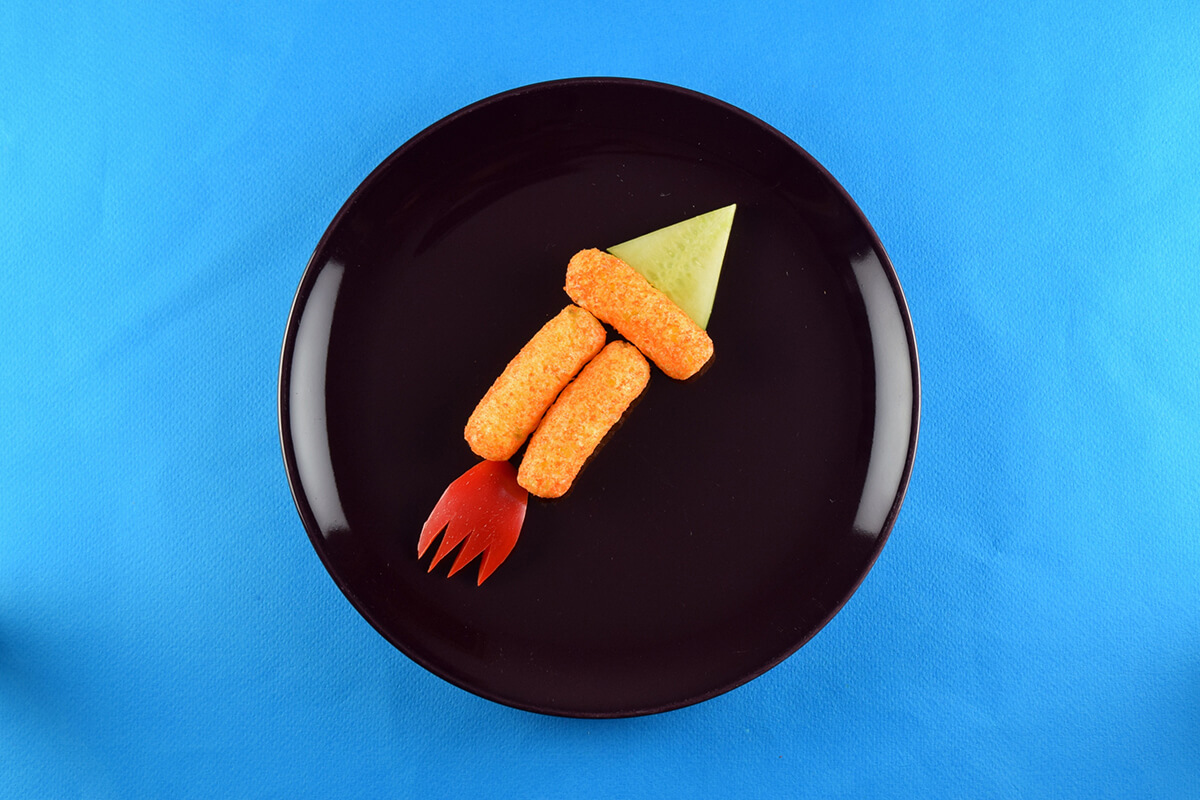 A triangle shaped cucumber slice is placed at the top of the rocket shape and a red pepper slice is cut in the shape of flames and placed at the bottom of the rocket shape
