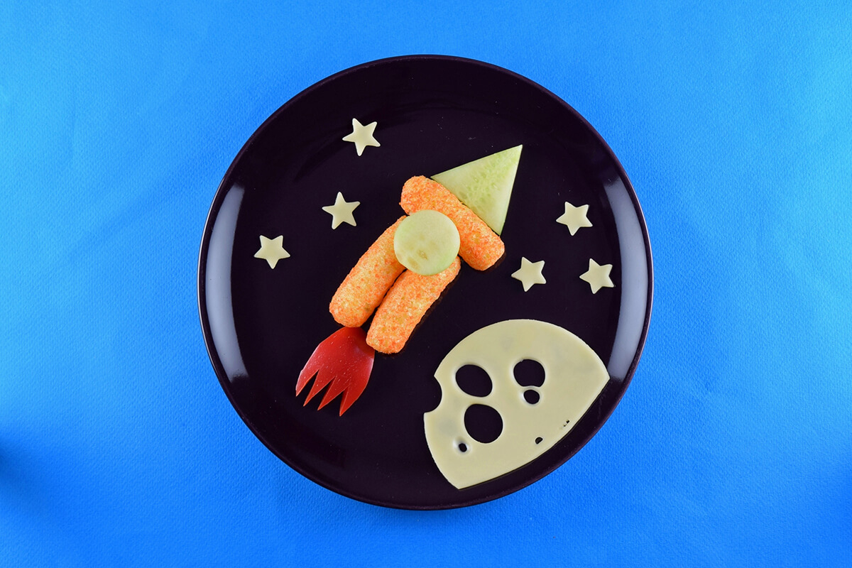 To complete the rocket, a circular cheese slice with holes cut into it is placed on the plate to create the moon and star shaped cheese stars are scattered around to create the night sky