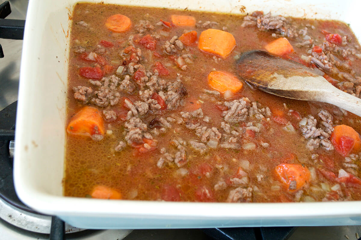 A casserole dish with cooked mince, finely chopped onion, carrots, pepper, herbs and tinned tomatoes and water