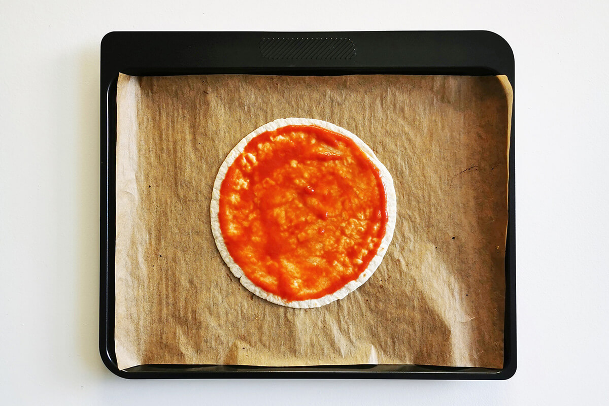 A lined baking tray with a tortilla wrap topped with pizza sauce in the centre