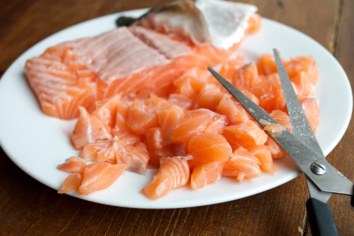A plate of raw salmon with the skin removed and some cut salmon pieces next to a pair of scissors
