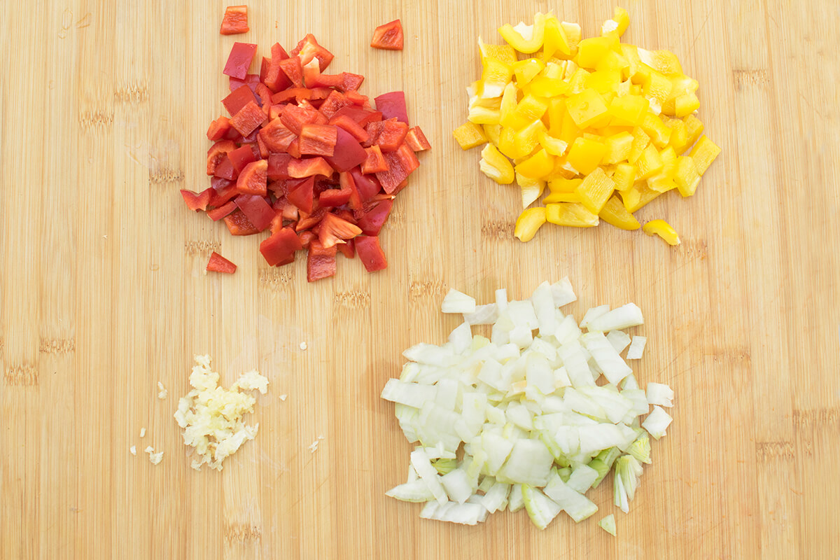 A chopping board with diced peppers, onion and a crushed clove of garlic 