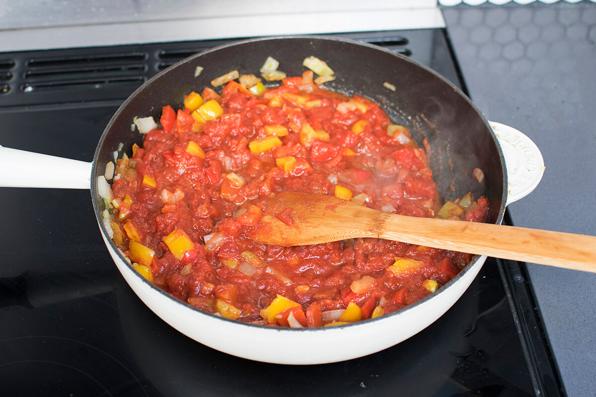 A frying pan of onion, garlic, peppers, herbs, spices, tomato paste and chopped tomatoes