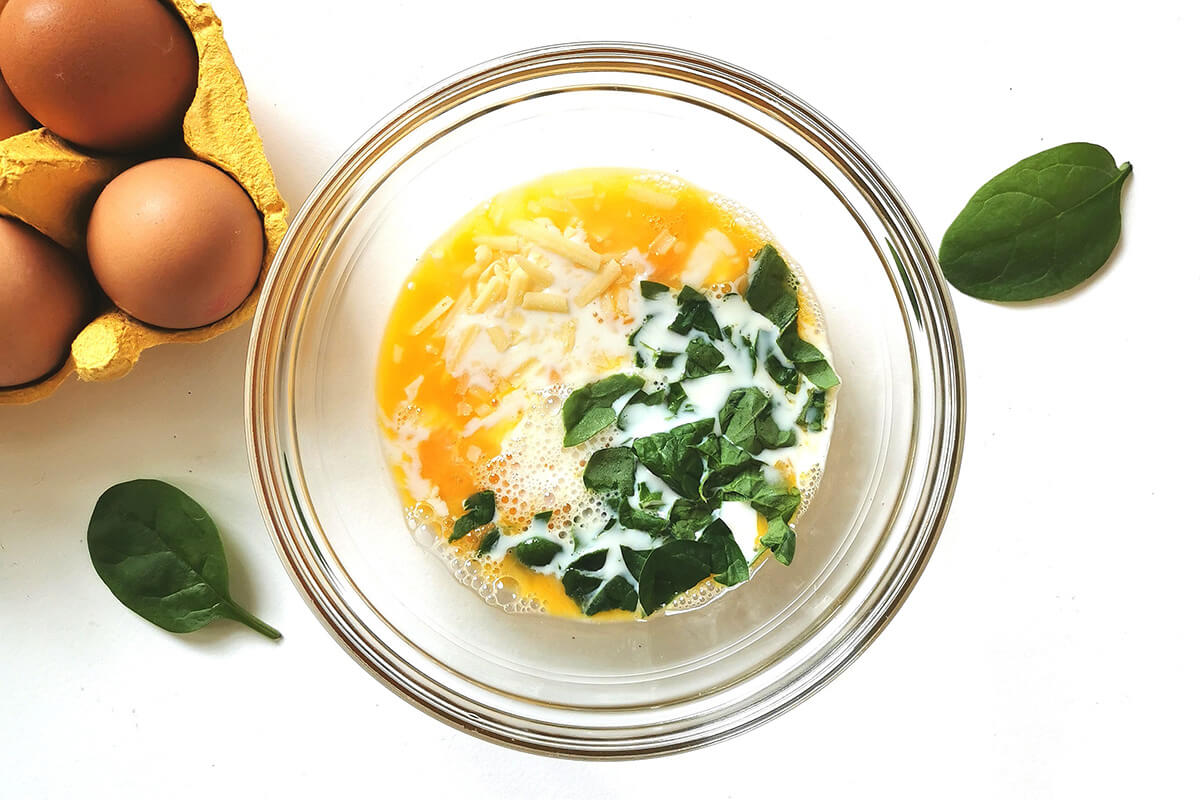 A bowl of whisked eggs with spinach and grated cheese, next to a crate of 4 eggs