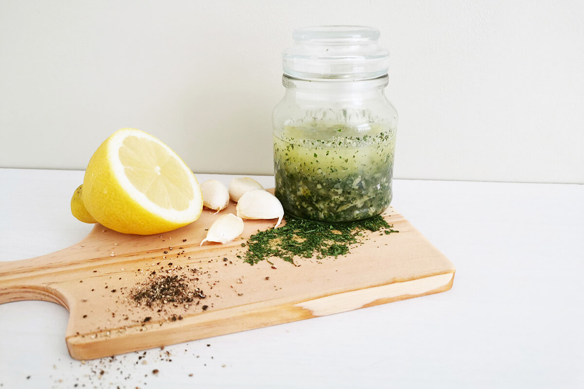 A chopping board with a jar of herby marinade, garlic cloves and half a lemon