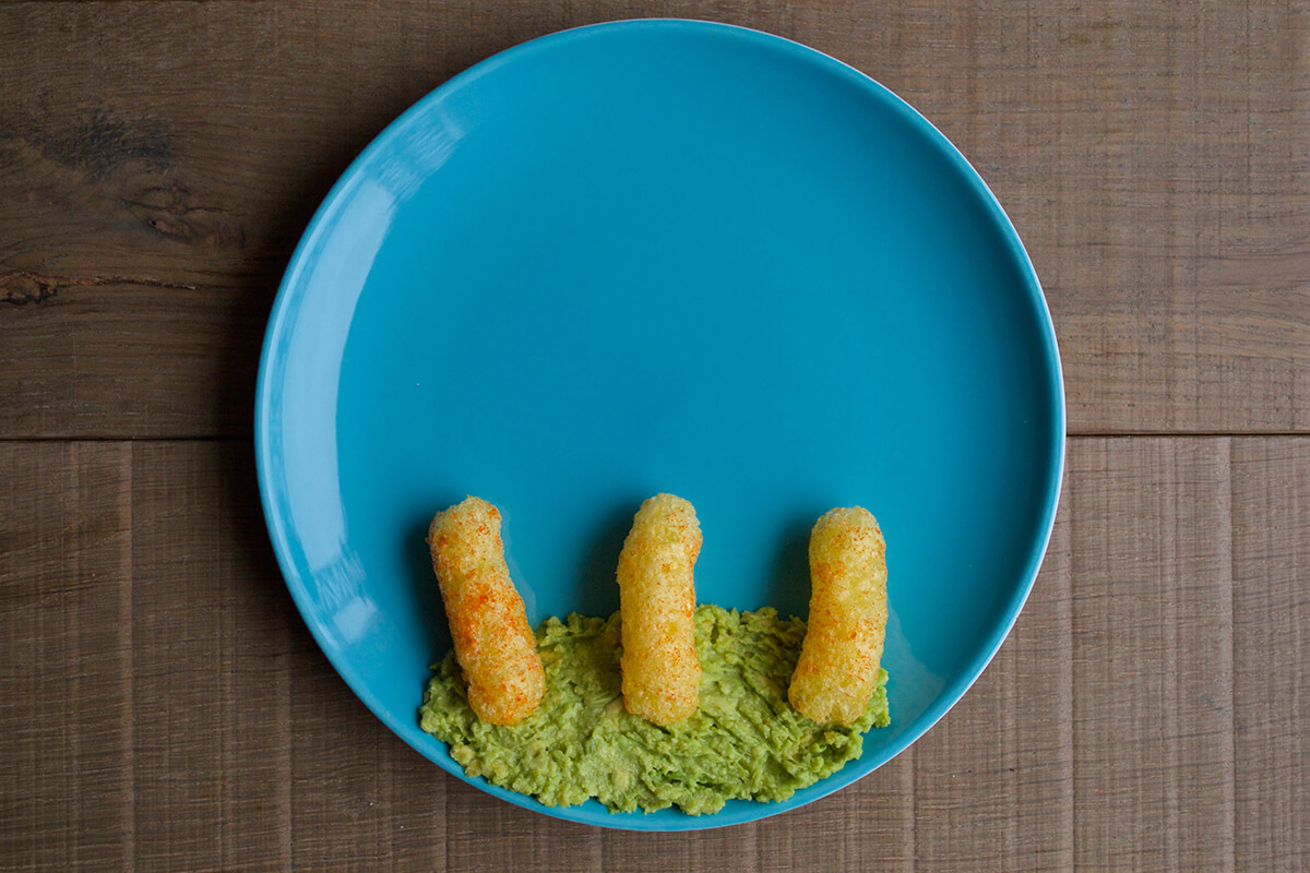 Mashed avocado is spread at the bottom of a plate with 3 organix melty carrot puffs arranged vertically on top of it