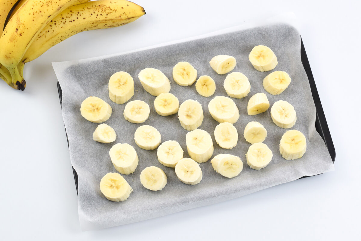 A baking tray lined with parchment paper with banana slices on it, next to a bunch of ripe bananas