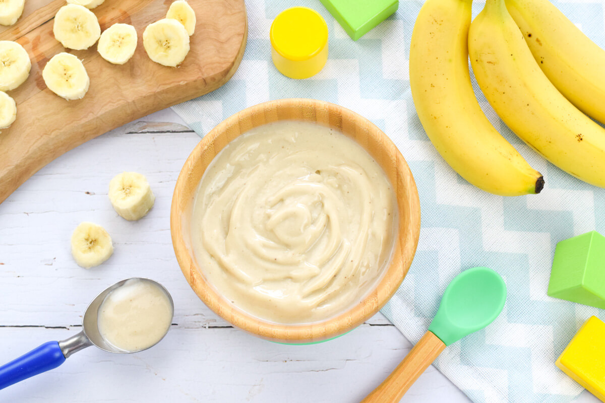 A bowl of vanilla nice cream next to a bunch of bananas and a wooden chopping board with sliced banana