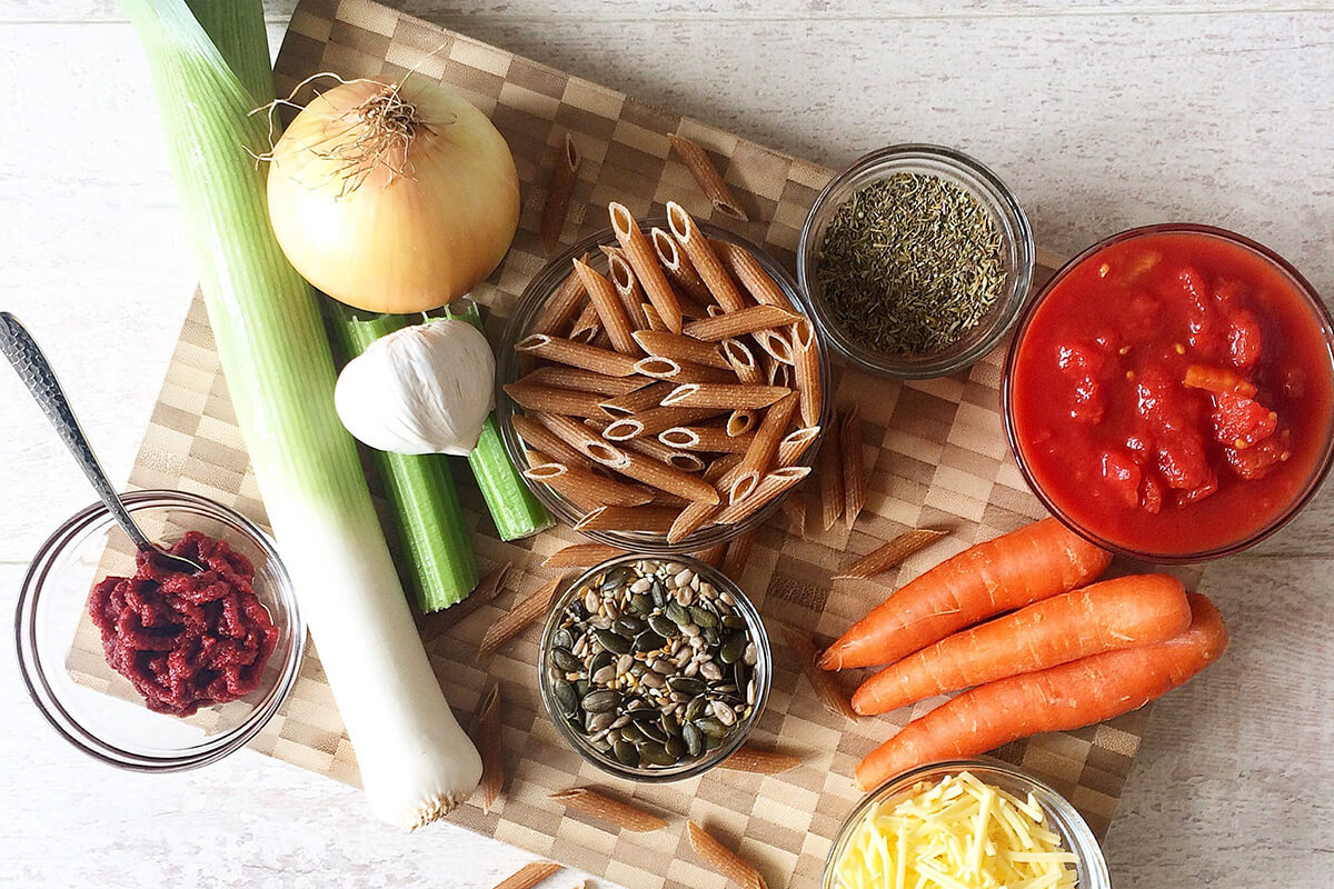 A chopping board with pasta, celery, leek, onion, carrots, chopped tomatoes, tomato puree, dried herbs and cheese