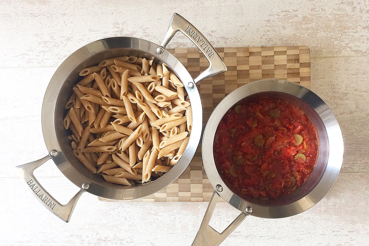 A chopping board with a saucepan of cooked pasta and a saucepan of vegetable tomato sauce mix on top of it