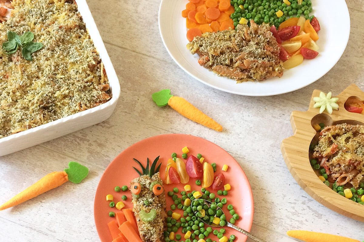 3 servings of vegetable pasta bake served with carrots, peas and sweetcorn, next to a casserole dish of pasta bake. One of the servings has a carrot shaped serving of vegetable pasta bake topped with carrots and cucumber to create eyes and pupils, a pea for a nose and a small celery slice for a mouth