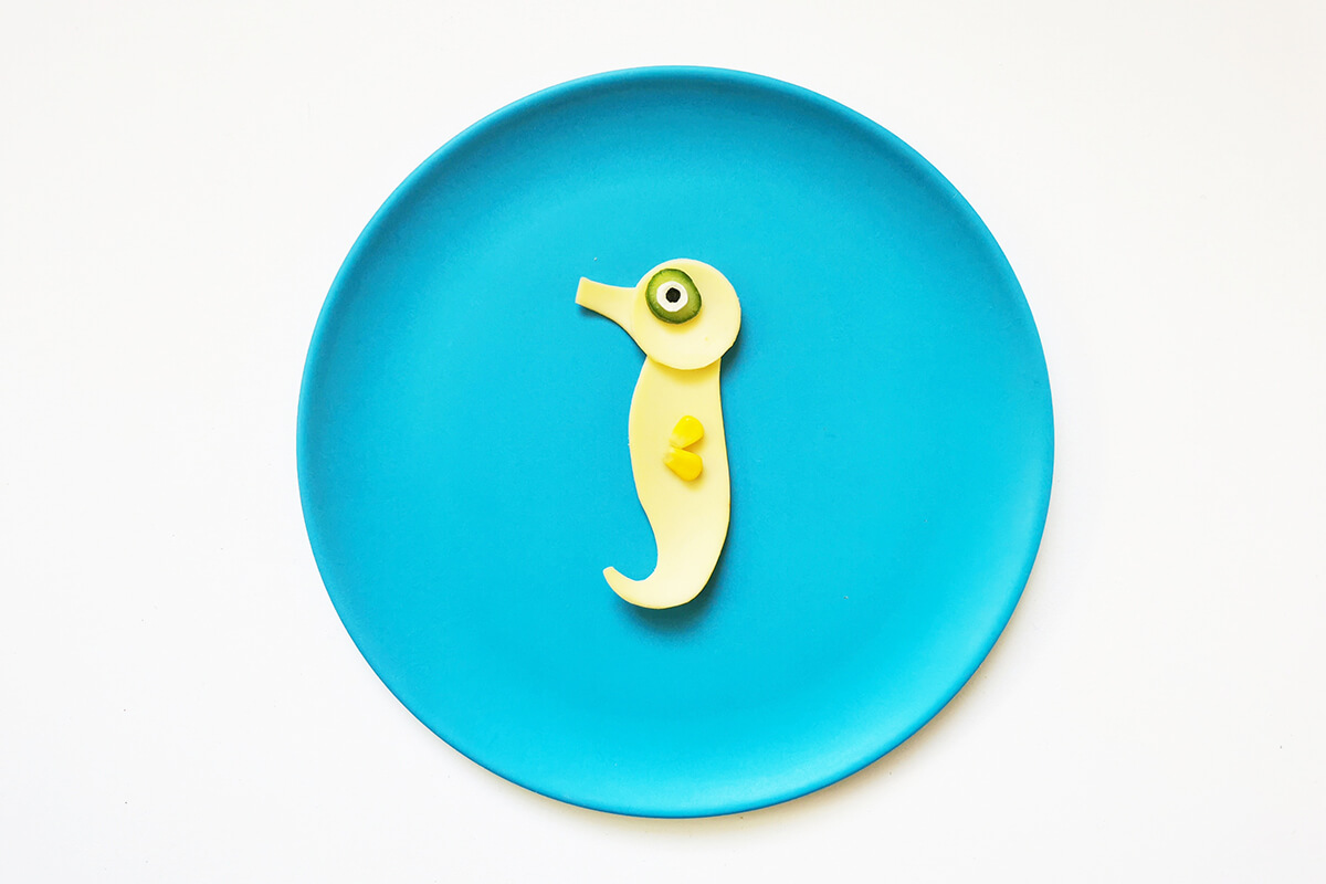 A mini cucumber circle is topped with a mini cheese circle and a pea and placed on the seahorse's head to create an eye. Two pieces of sweetcorn are added to create the fin