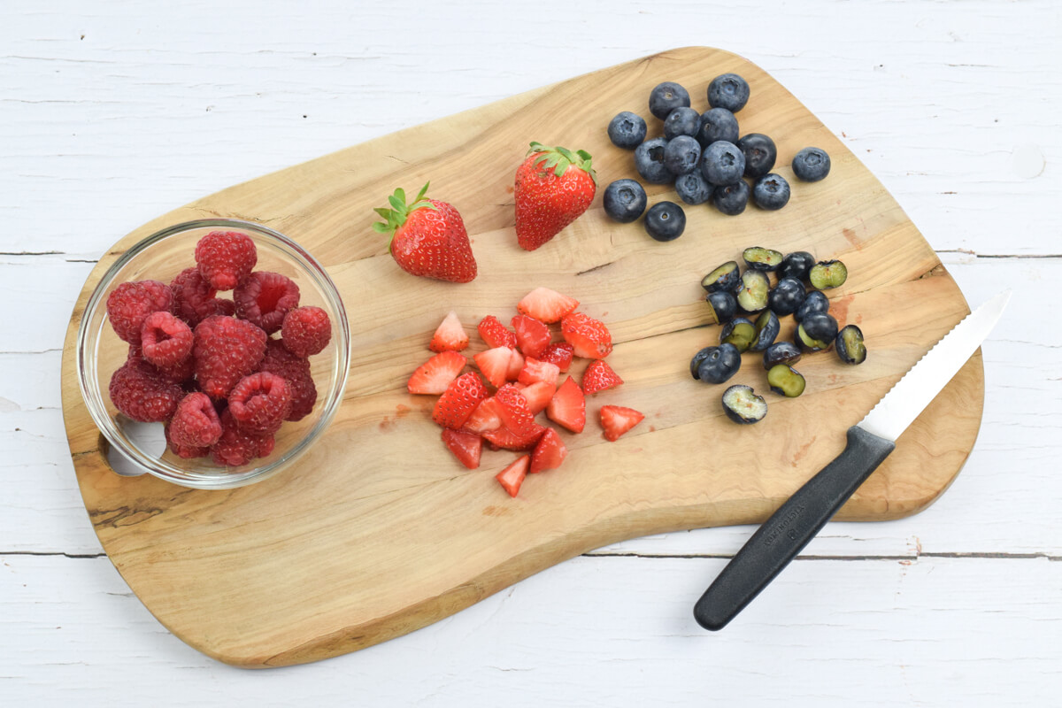 A wooden chopping board with some whole and chopped blueberries and strawberries, and a small glass bowl of raspberries