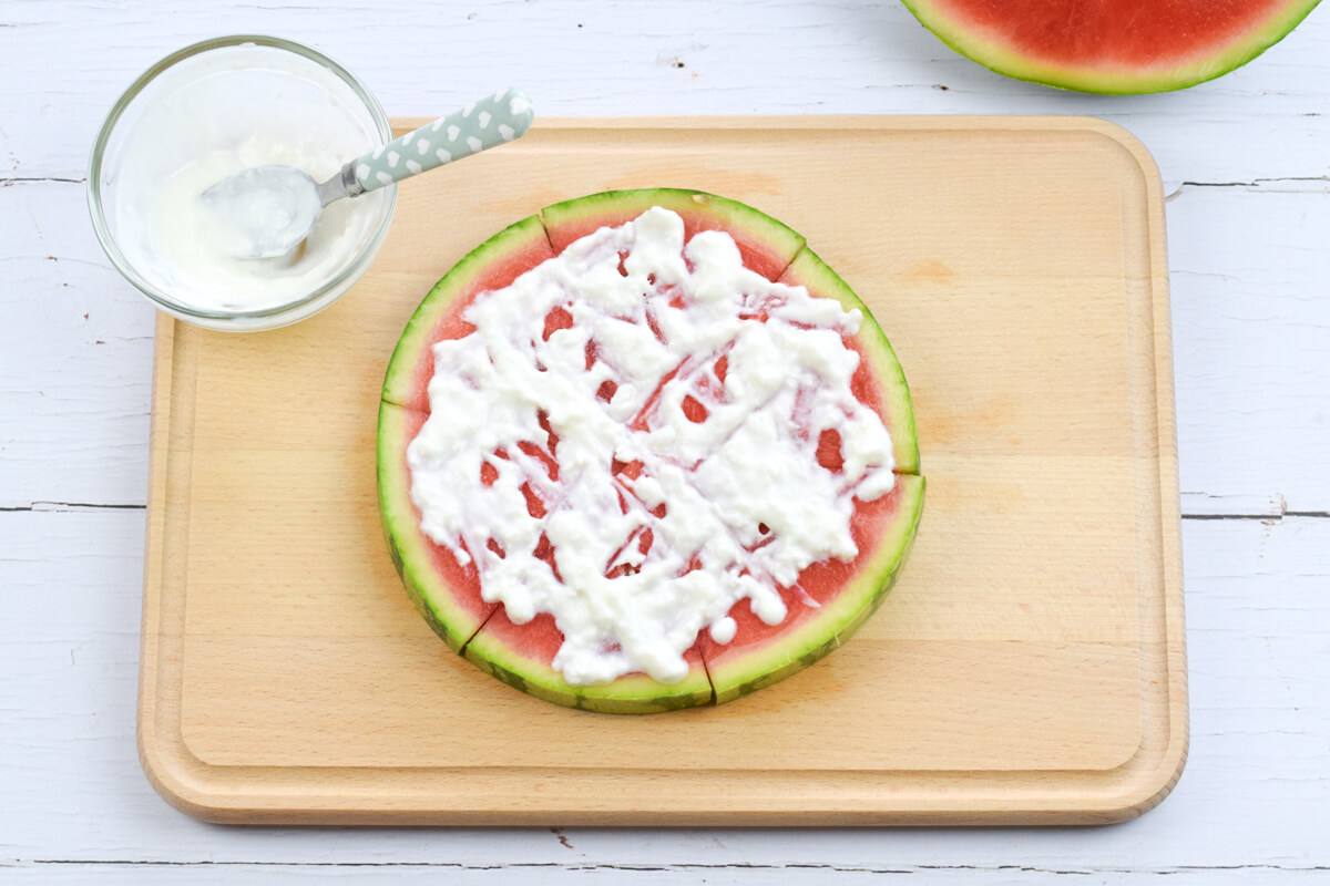 A wooden chopping board with a watermelon round cut into 6 triangle shaped segments and topped with yoghurt