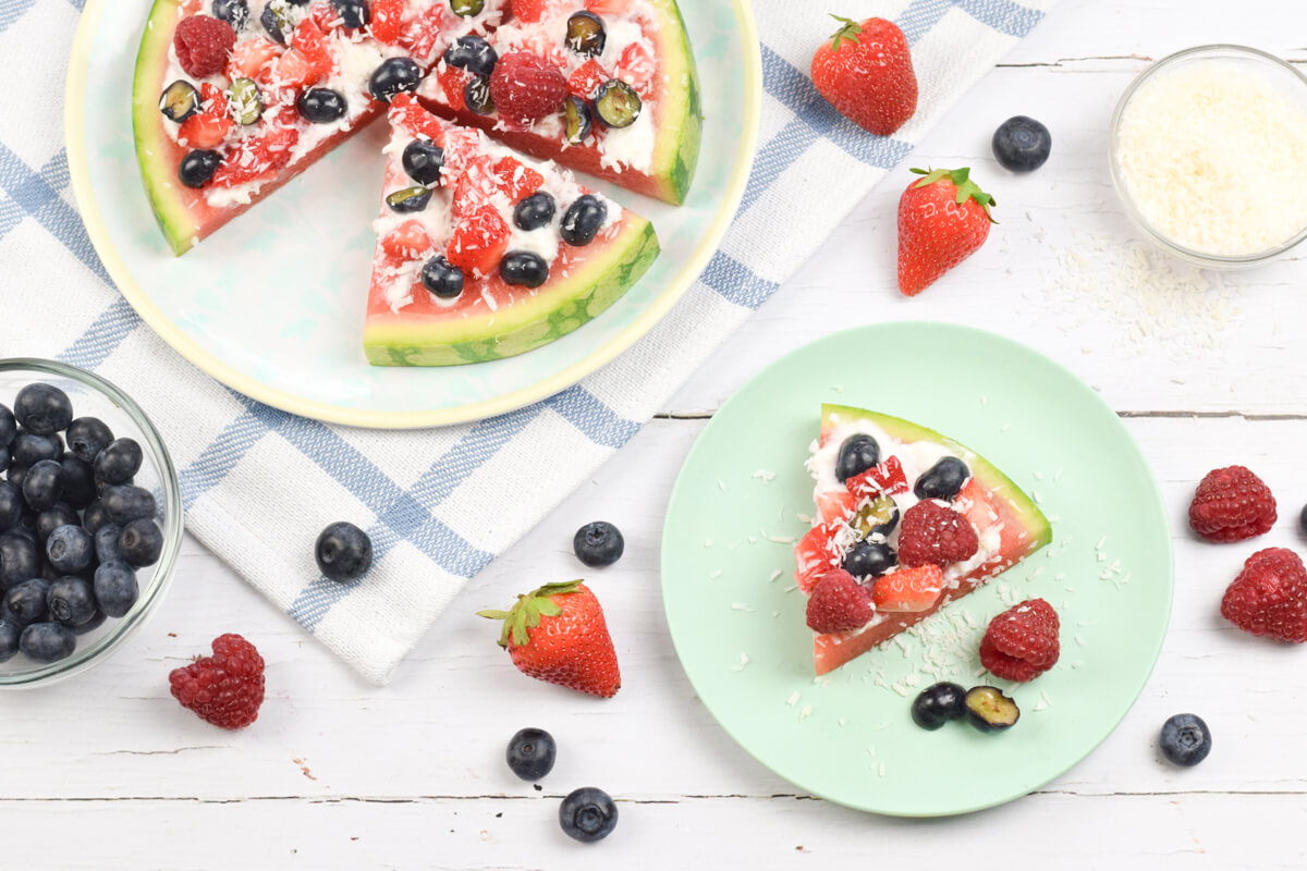 A serving of watermelon pizza next to a plate of watermelon pizza slices topped with yoghurt, blueberries, raspberries, strawberries and desiccated coconut 