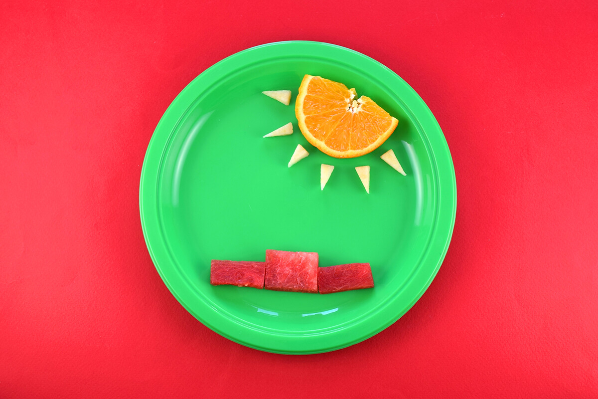 An orange slice is placed at the top of the plate and apple triangles are placed coming off it to create a sun with sun rays