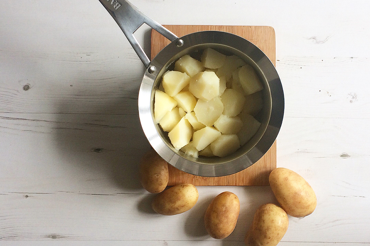 A saucepan of boiled potatoes on a shopping board with some whole potatoes next to it