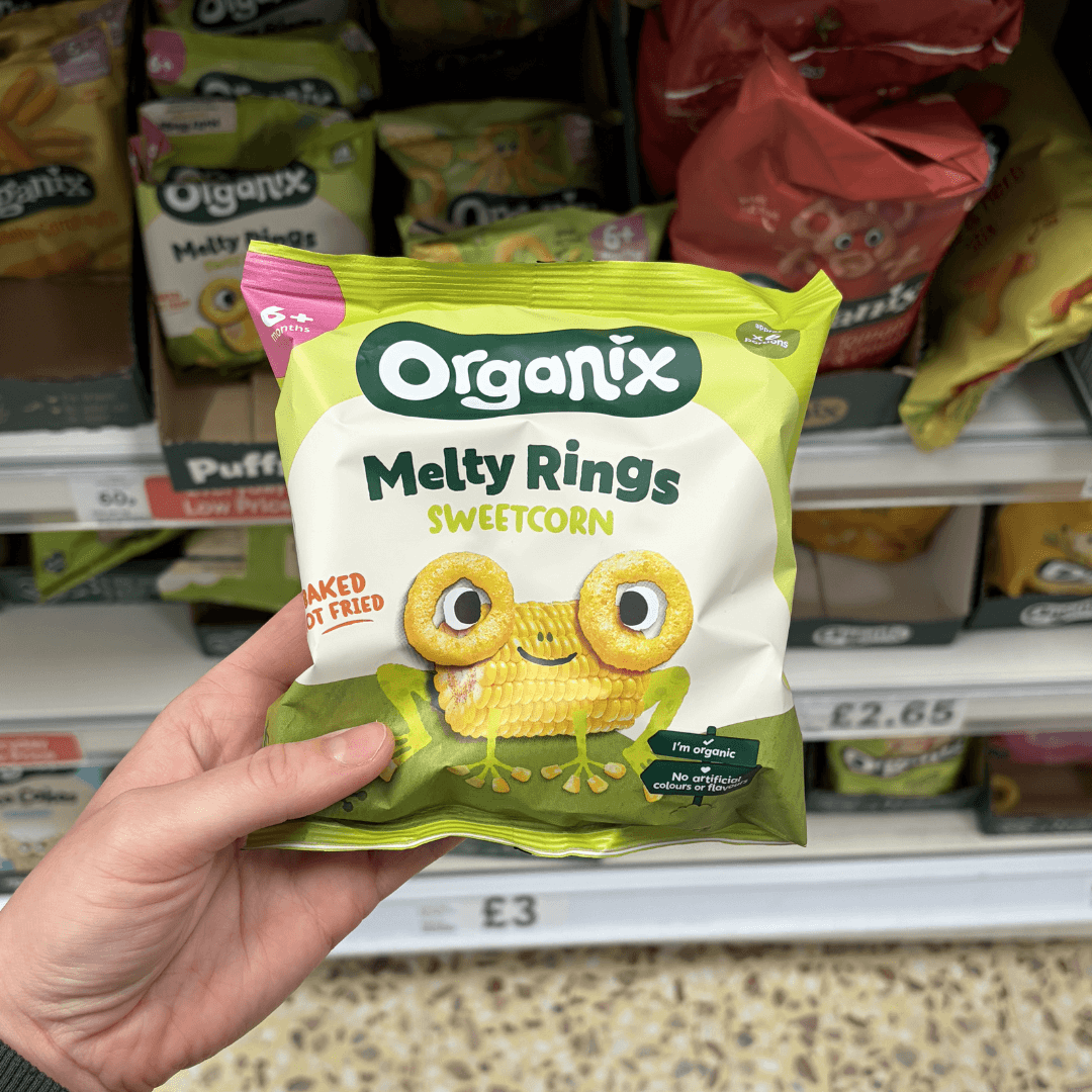 Someone holding a new organix sweetcorn rings packet in the supermarket in the baby food isle