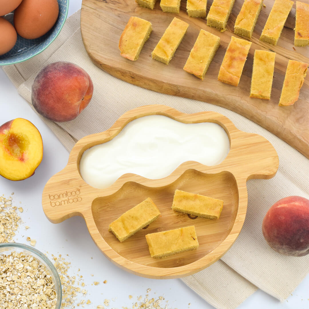 A serving of peach baked oat fingers with yoghurt, next to a chopping board of peach baked oat finger slices and some peaches