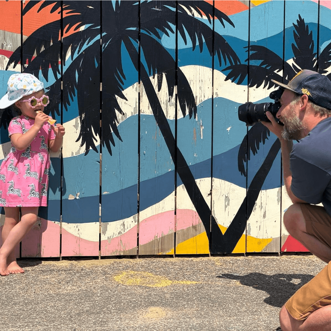 Toddler dressed in pink dress wearing a sun hat and sun glasses and eating an Organix oaty bar while leaning on a summery wall with a the image of a beach. An adult is taking a picture of the toddler.