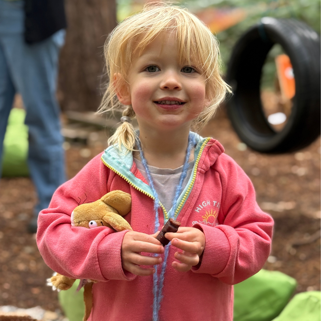 A sweet toddler wearing a red jacket, and holding a little mouse plush toy from the Gruffalo story, as well as an Organix fruit bar. They are smilign to the camera. 